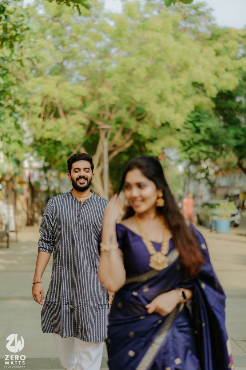 A couple in traditional Indian attire pose in a park setting with green trees in the background.