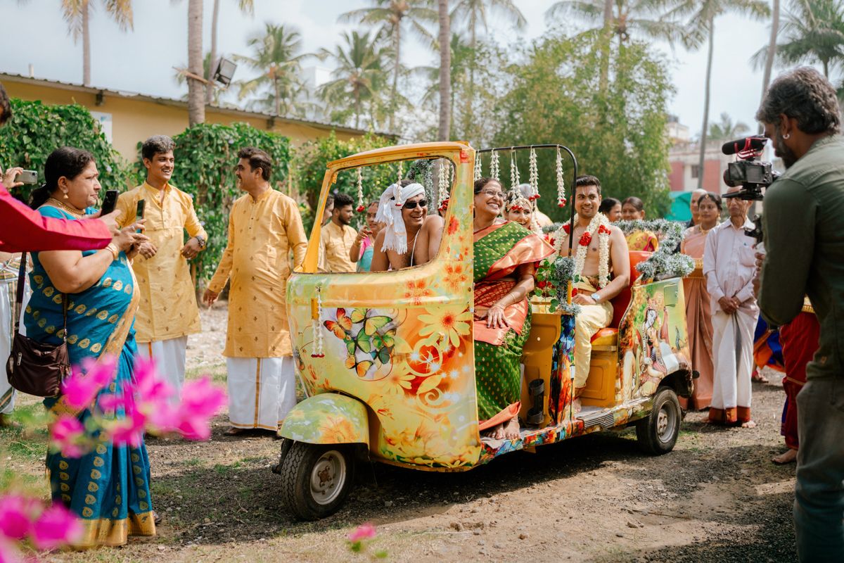 A decorated auto rickshaw with yellow and floral decorations surrounded by guests at an Indian wedding celebration.