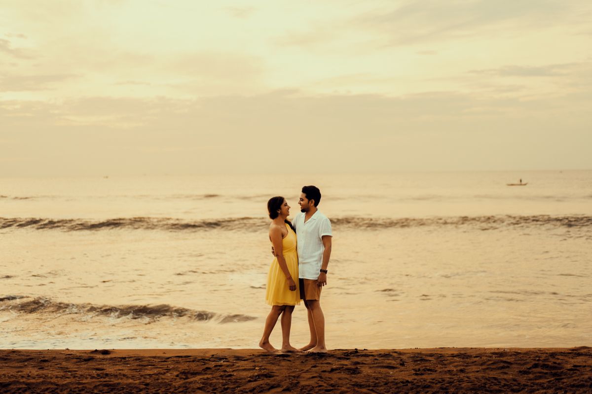 A couple walks along the shoreline at sunset, silhouetted against a golden sky and calm ocean waves.