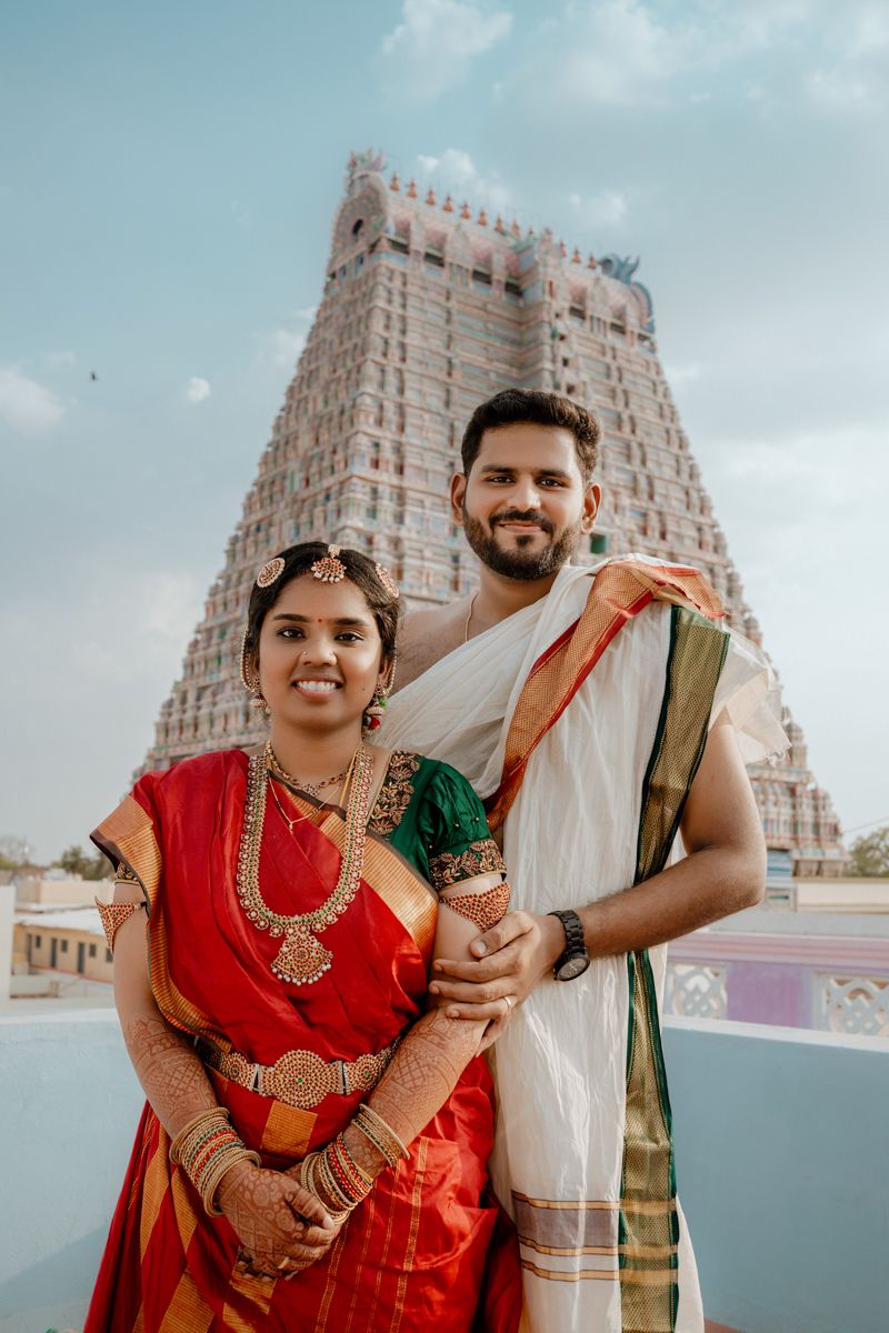 A couple in traditional South Indian attire poses in front of a grand temple gopuram tower on a sunny day.
