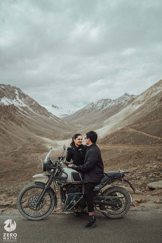 Vintage motorcycle parked along mountainous road with dramatic rocky peaks and cloudy sky in background.