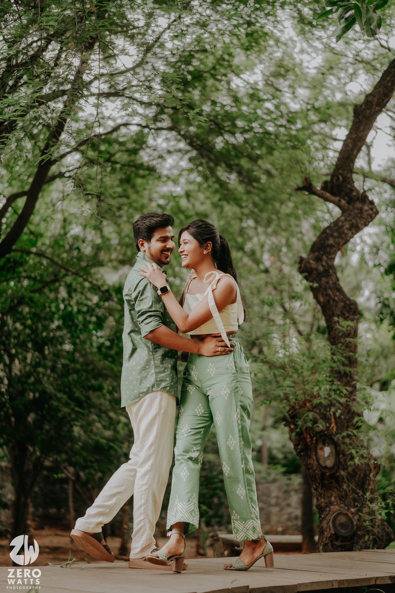 A couple shares a romantic moment while embracing on a wooden deck surrounded by lush green trees in a natural setting.