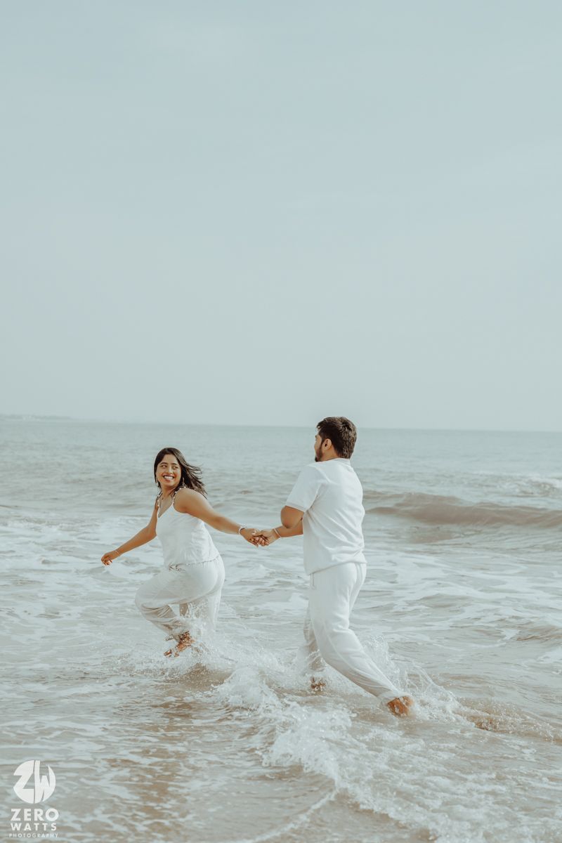 Joyful couple running through ocean waves in white outfits.