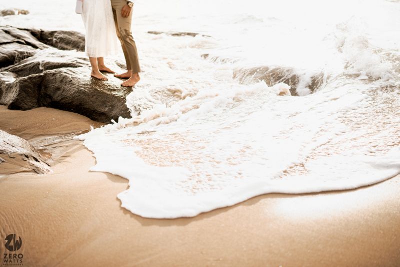 Waves crash against rocks as feet stand at the edge of sandy beach.