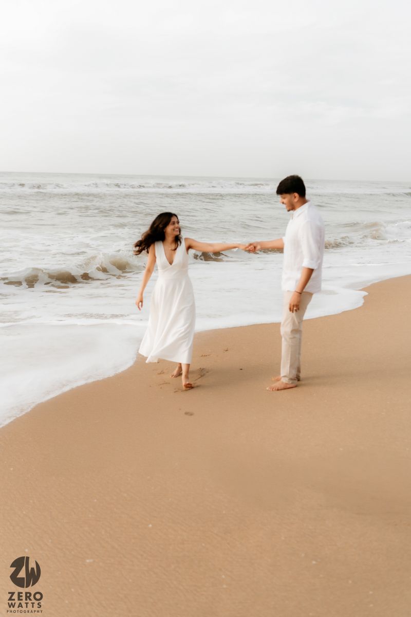 A couple holds hands at the edge of the ocean waves wearing matching white outfits.