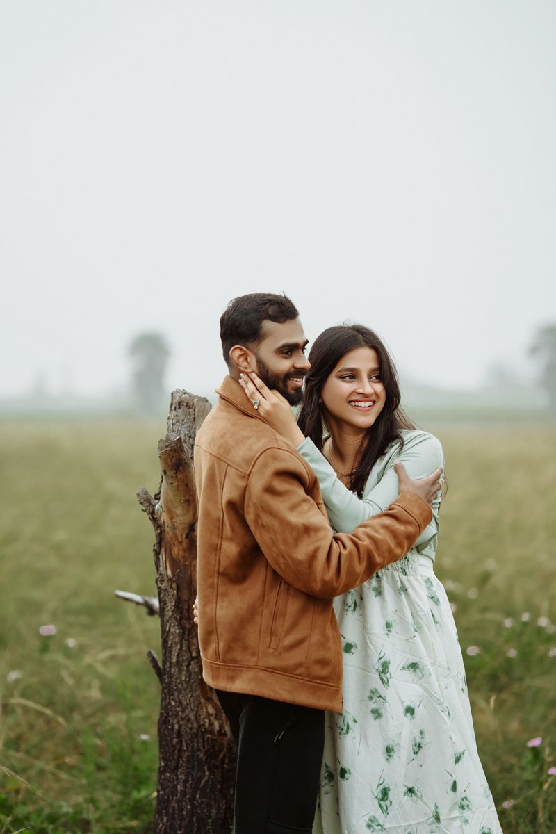 A couple embraces and smiles while standing beside a rustic tree trunk in a meadow.