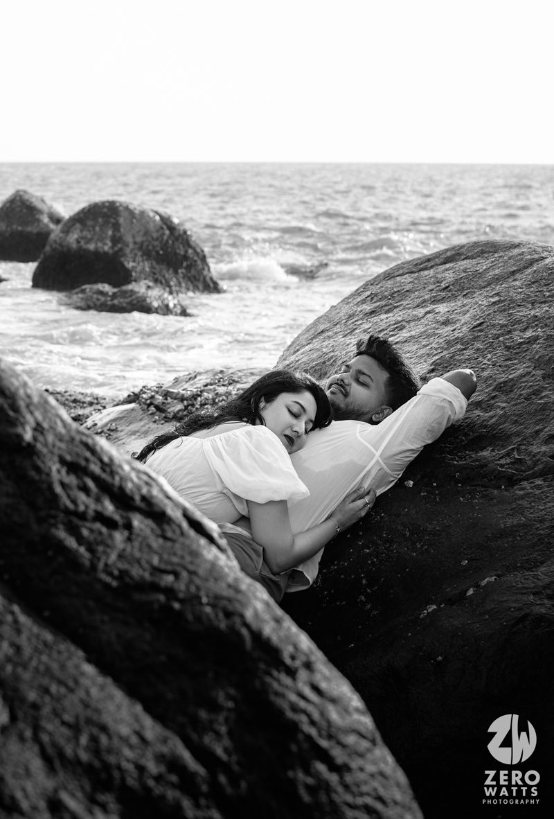 A couple rests intimately against coastal rocks as waves crash in the background.