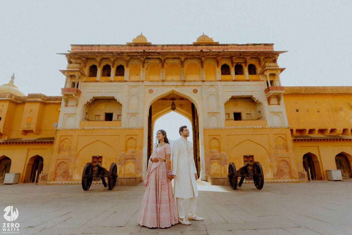 A couple in traditional attire stands in front of the majestic Amer Fort in Jaipur, India, with two elephant statues flanking them.