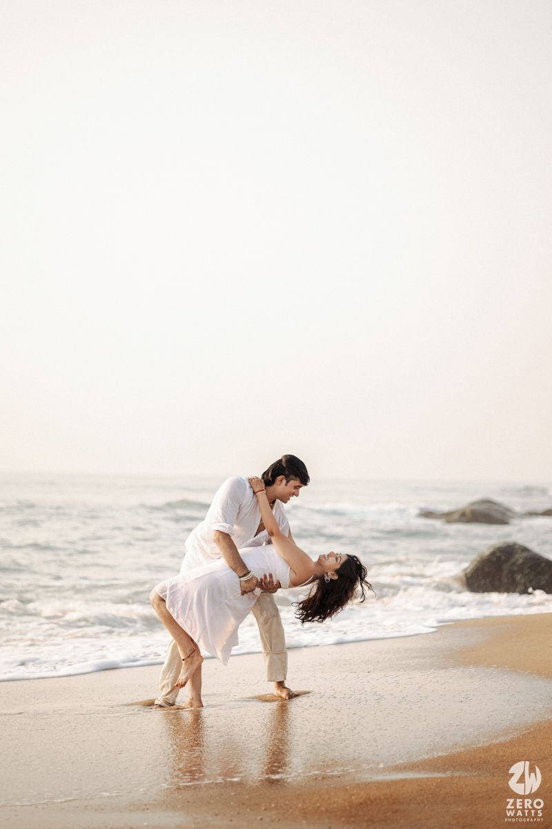 Couple in white outfits dances together on sandy beach with ocean waves and rocks in background.
