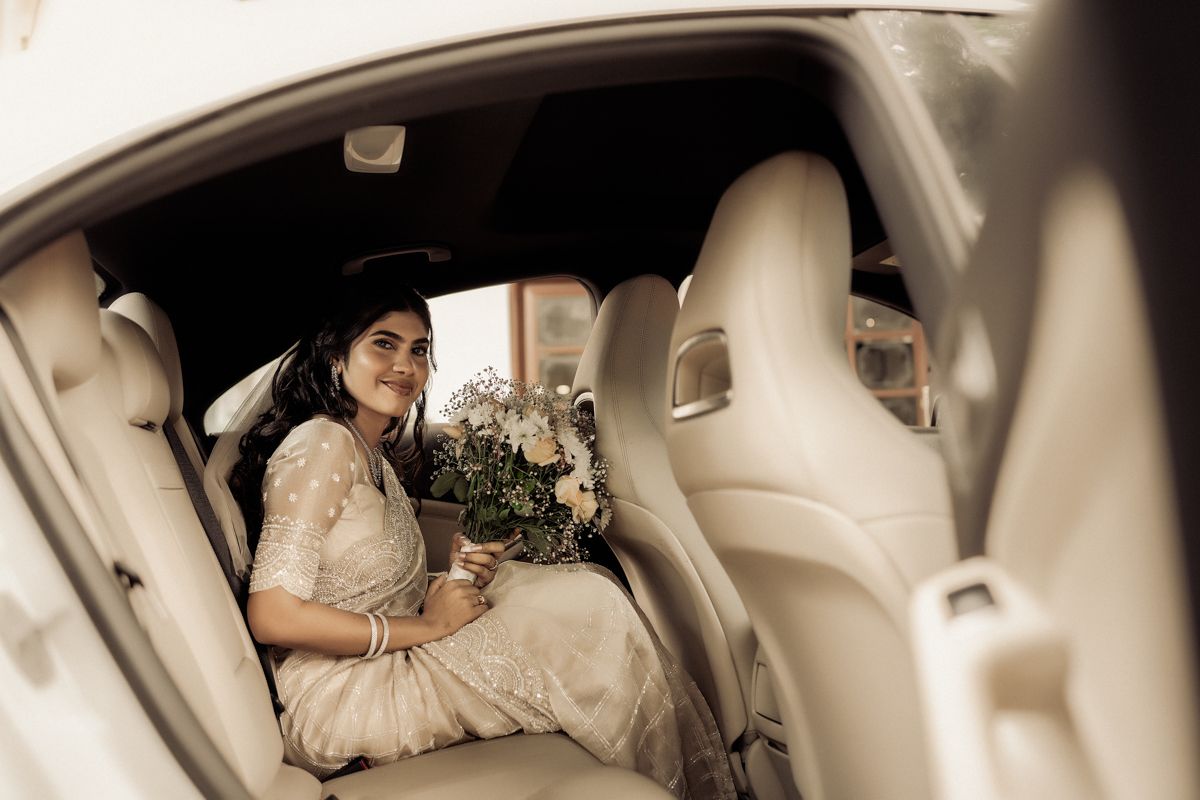 Elegant bride in cream-colored dress sits in luxury car backseat holding wedding bouquet in sepia-toned photo.