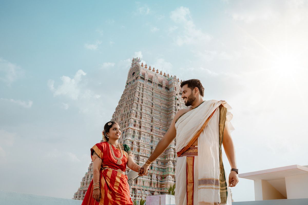 Two people in traditional South Indian attire stand before the towering Meenakshi Temple gopuram in Madurai.