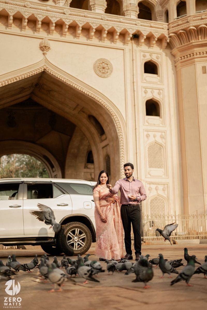 Couple poses in front of ornate Indian architecture with a white SUV and pigeons in the foreground.