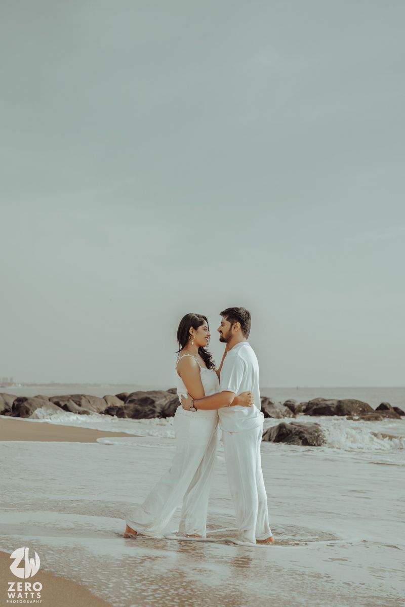 Couple in white outfits embracing on a rocky beach during an overcast day.