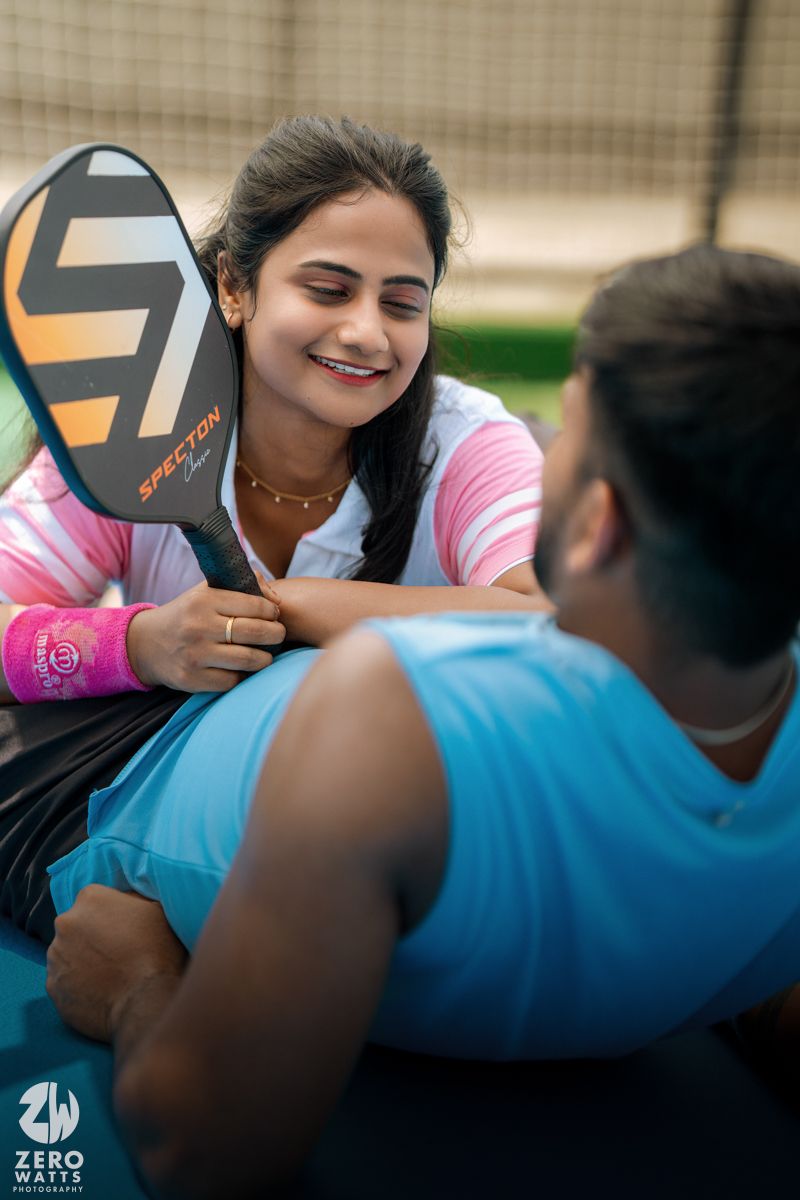 Playful couple lying on tennis court sharing a lighthearted moment with a paddle.