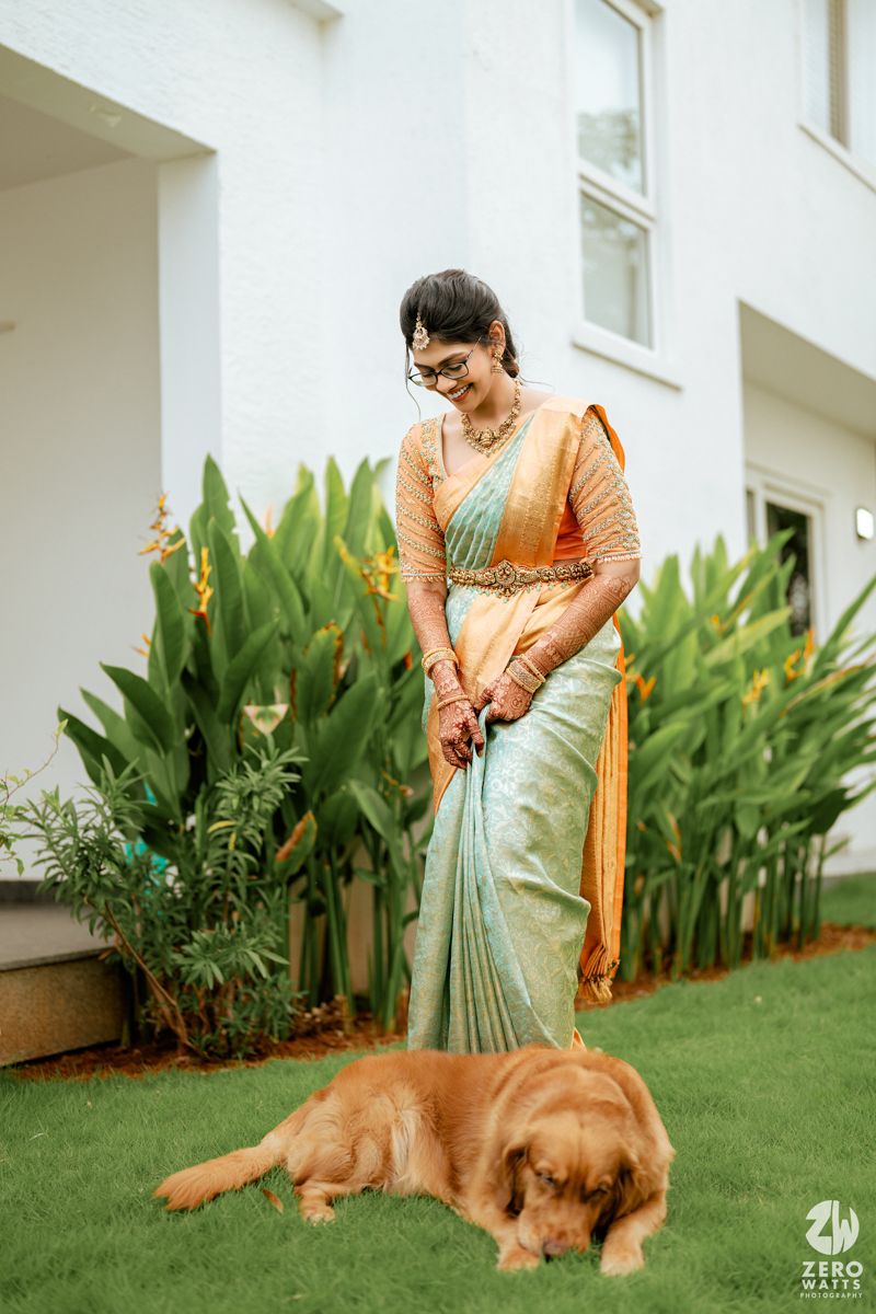 A woman in a mint green and orange sari stands on a lawn with a resting golden retriever dog beside her.