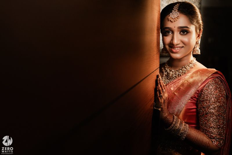 Beautiful bride wearing traditional Indian jewelry and red saree poses elegantly against dark background with soft lighting.
