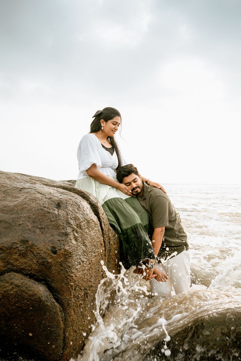 Two people sharing an intimate moment on coastal rocks as ocean waves splash around them.