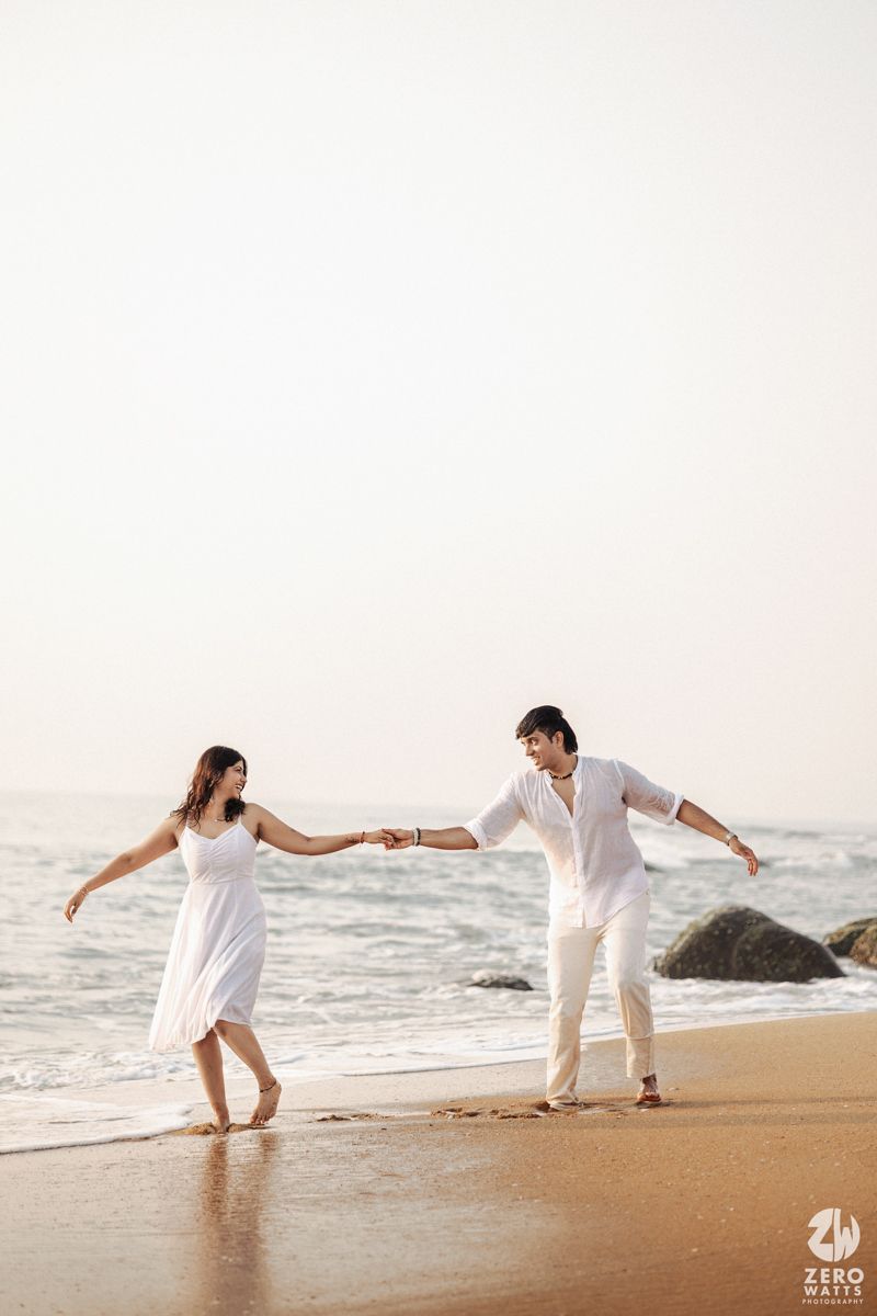 A romantic couple holds hands while dancing barefoot on a sandy beach with ocean waves and rocks nearby.