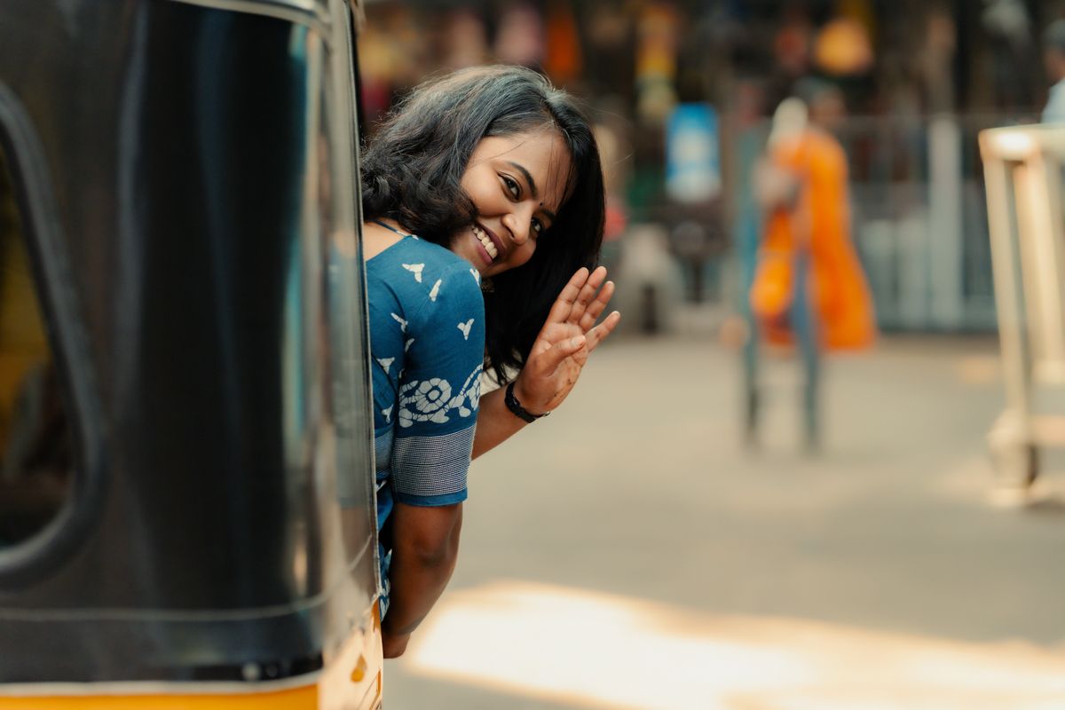 Person waving from yellow auto rickshaw window with joyful expression on sunny street in India.