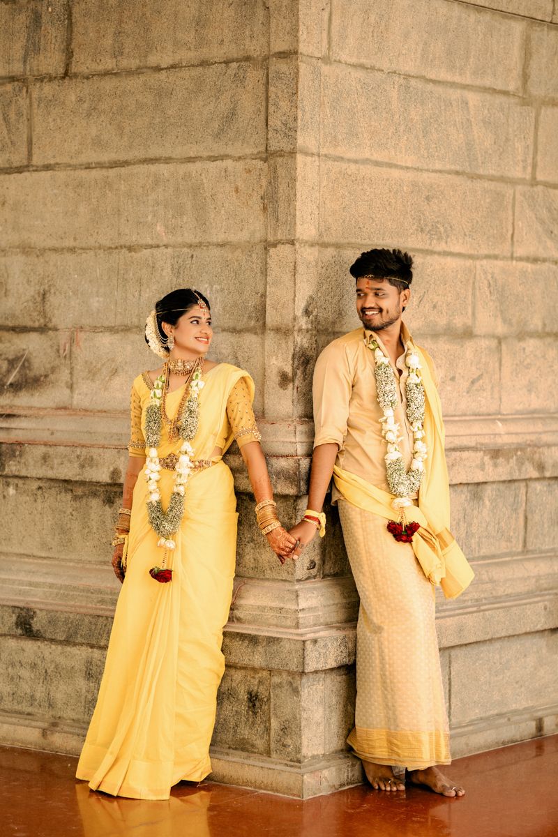 A couple in matching yellow traditional South Indian attire holding hands and posing against a stone wall.