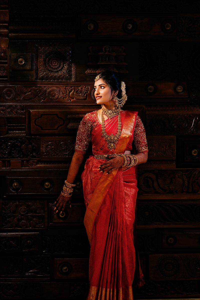 Traditional South Indian bride wearing red silk saree with gold jewelry stands against ornate wooden carved wall.