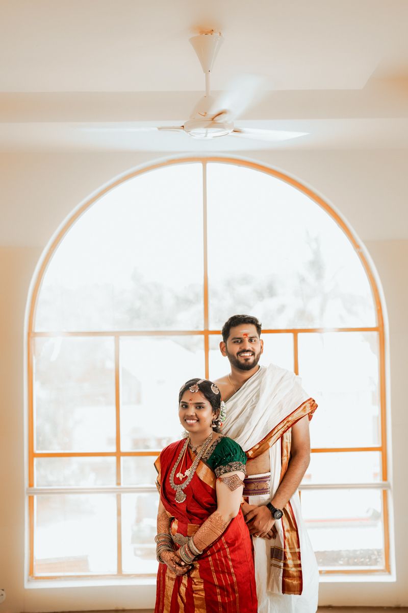 Two people wearing traditional Indian attire stand together in front of an arched window with soft natural lighting.