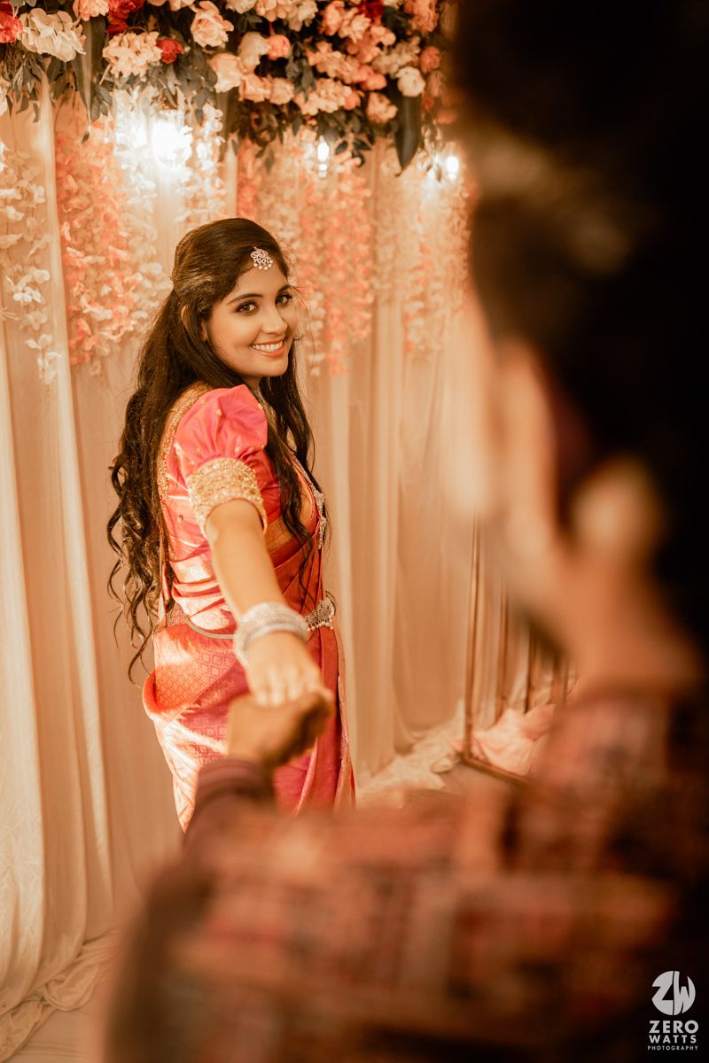 A person in a pink silk saree poses gracefully against a floral decorated backdrop at an indoor celebration.