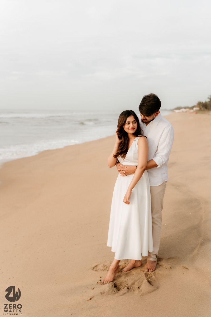 A romantic couple embraces on a misty beach wearing white and beige clothing.