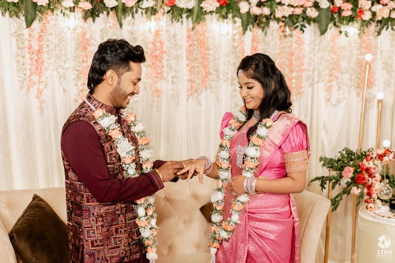 Couple in traditional Indian attire exchange rings during engagement ceremony with floral backdrop and decorations.