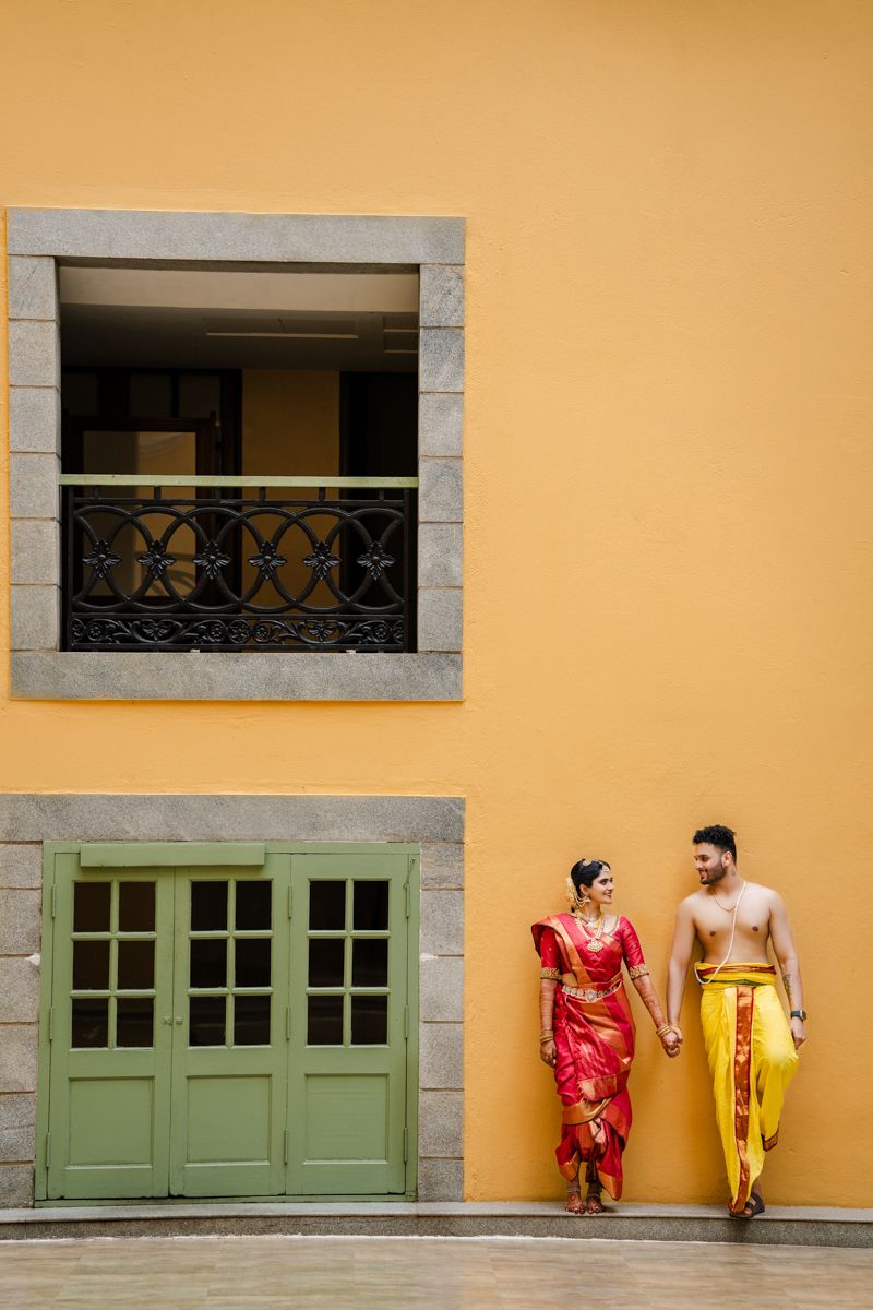 Two people in vibrant traditional attire pose against a yellow wall with green doors.