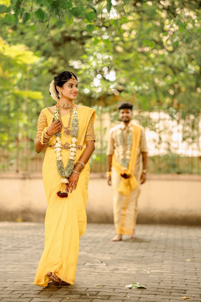 A couple in matching yellow traditional Indian outfits adorned with flower garlands pose outdoors in a garden setting.