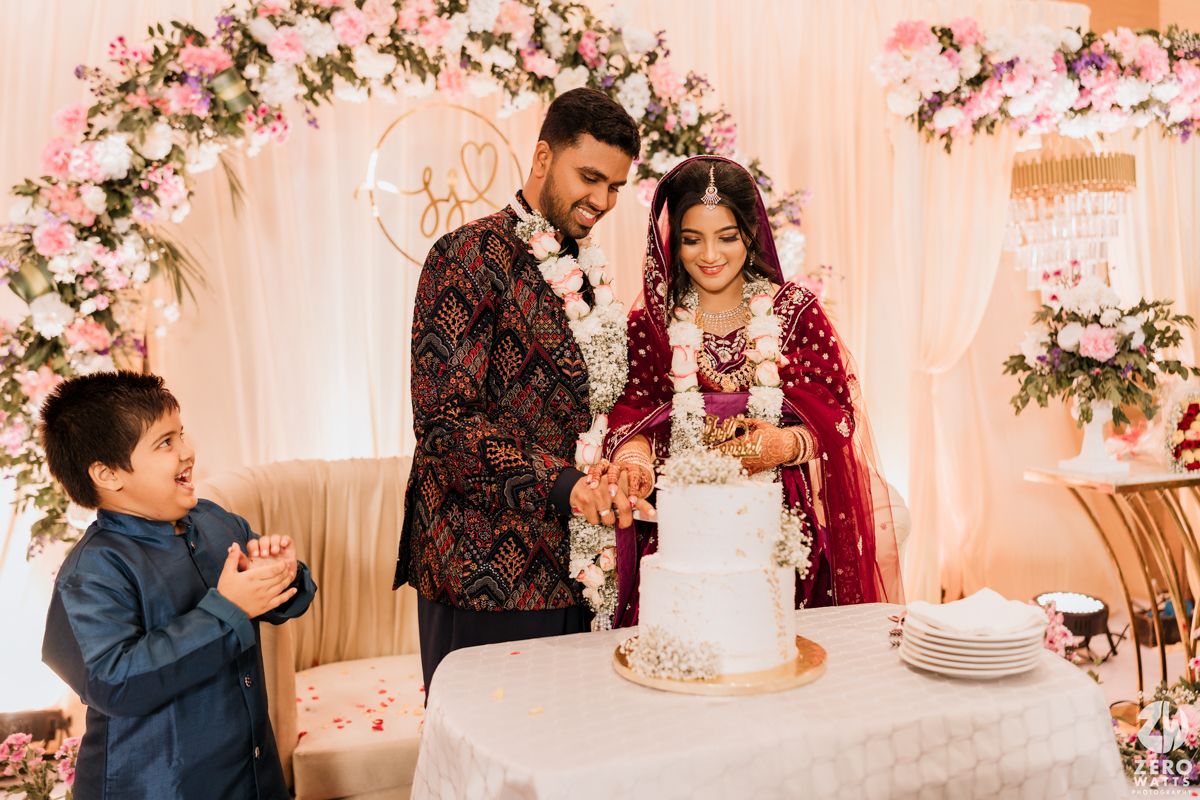 Couple cutting their wedding cake together under floral arch decoration while young child watches.