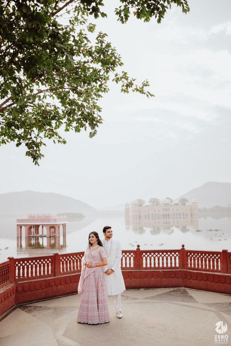 A couple poses on a red-railed terrace with the Jal Mahal palace visible across the lake in Udaipur.