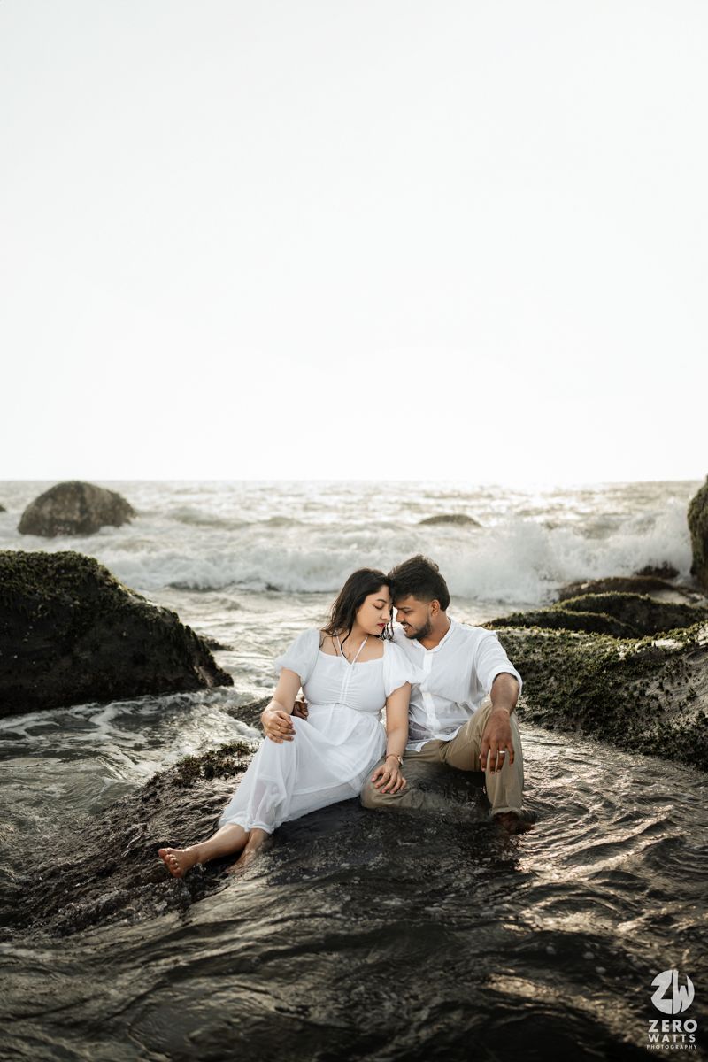 A couple in white outfits shares an intimate moment while sitting on rocky ocean shore.