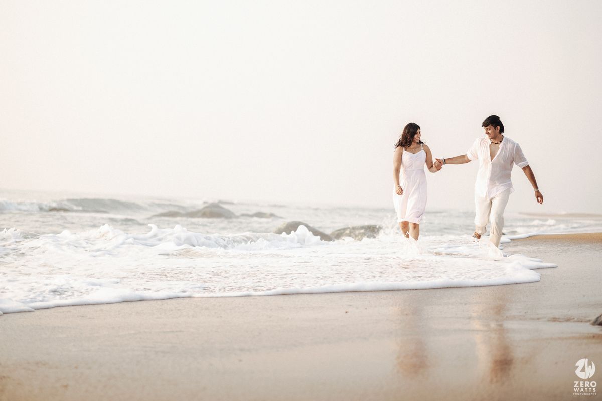 Couple in white clothing holding hands and playing in the ocean waves on a sandy beach.