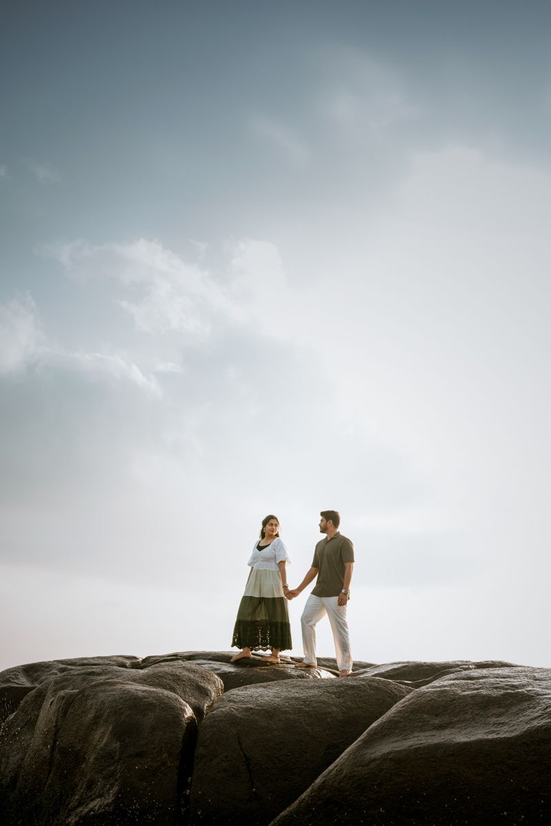 Two figures stand together on rocky coastal cliffs against a bright, cloudy sky at sunset.