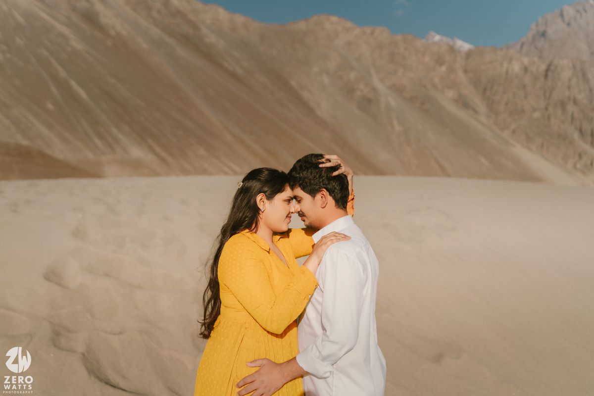 Couple in white and yellow outfits embrace with rocky desert mountains serving as dramatic backdrop at sunset.
