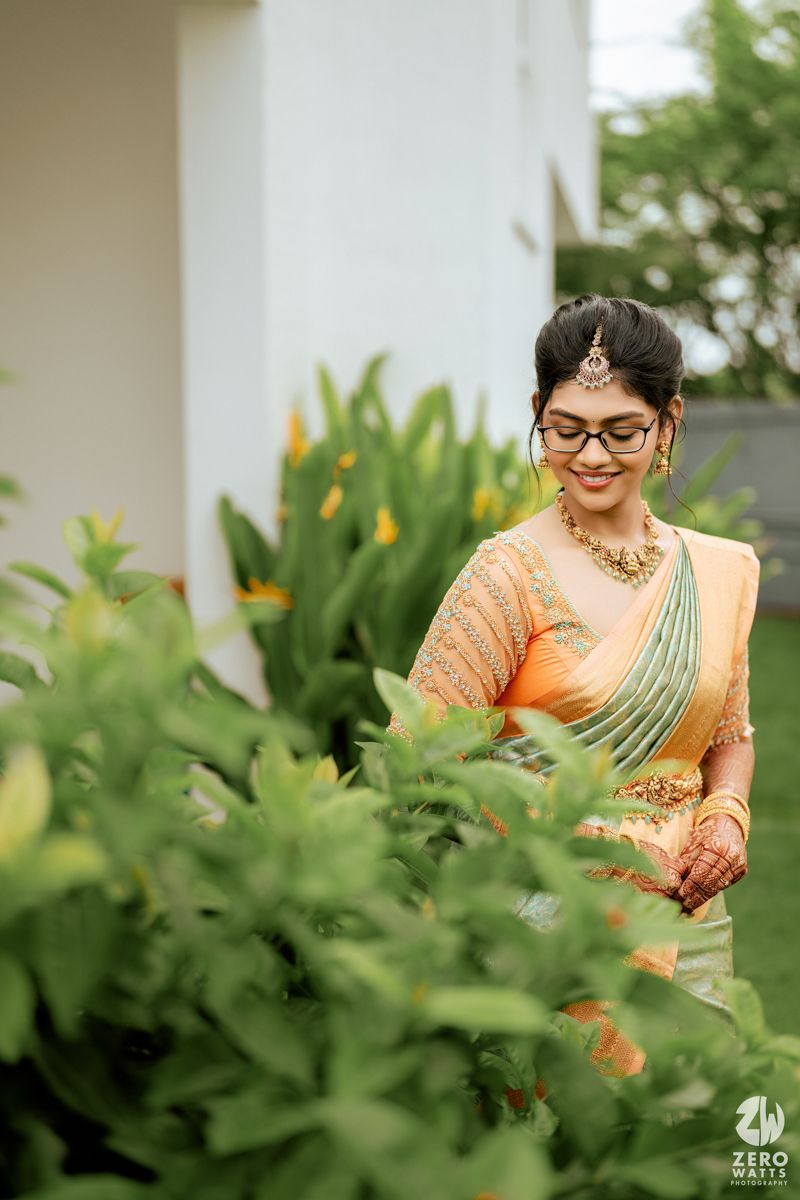 A person in a mint green and orange sari with glasses poses elegantly among lush greenery in an outdoor setting.
