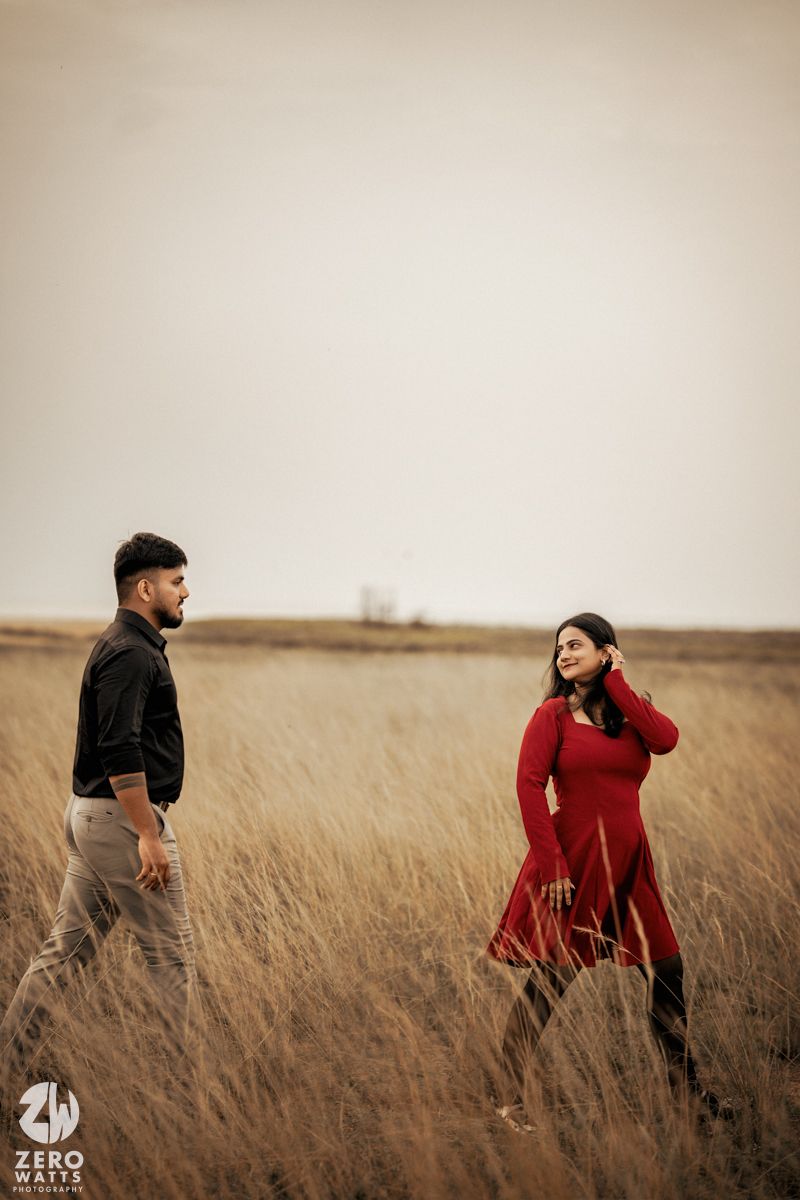 Romantic couple walking through golden wheat field at sunset wearing red dress and dark clothing.