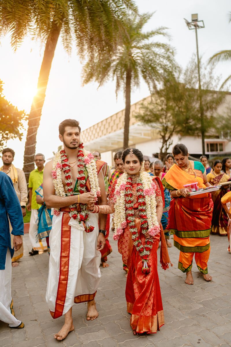 Traditional Indian wedding procession outdoors at sunset with palm trees, featuring couple in red floral garlands and formal attire.