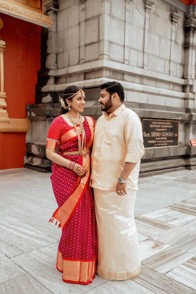 A couple dressed in traditional Indian wedding attire stands at the temple entrance wearing red and cream colored outfits.