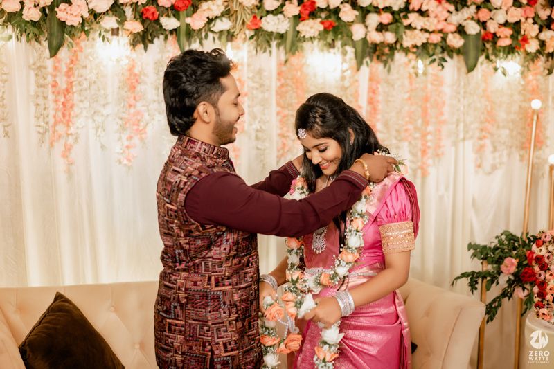 A couple in traditional Indian attire sharing a sweet moment at an event with floral decorations and lighting.