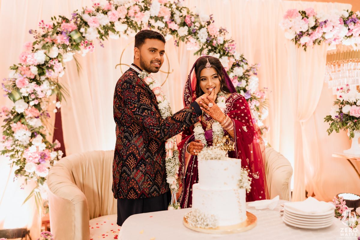 Newlyweds cutting their elegant white wedding cake under a decorative floral arch.