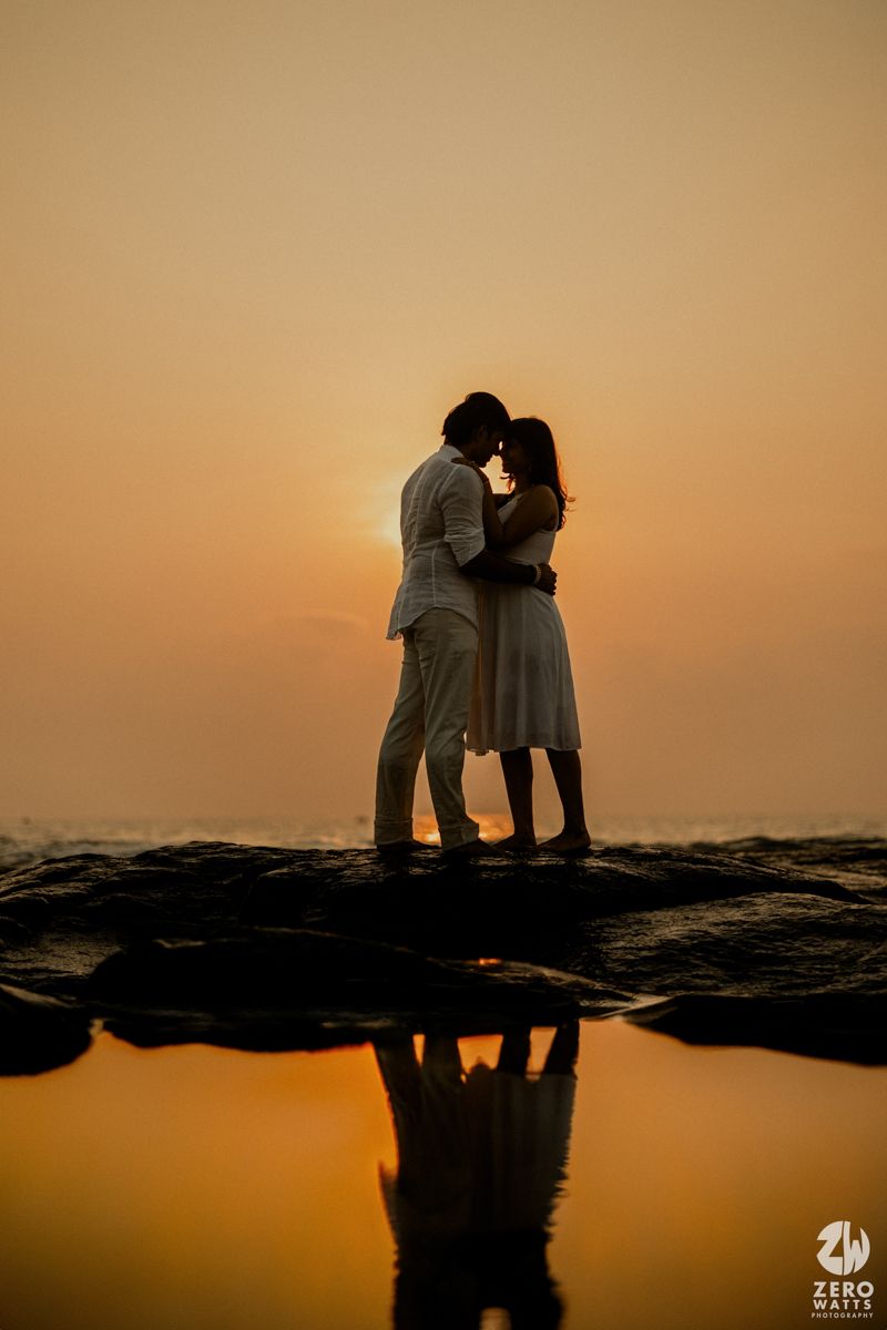 Silhouetted couple standing on coastal rocks at sunset with perfect reflection in tidal pool.