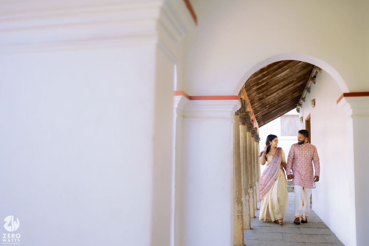Two people in traditional attire walk down a white-walled corridor with an arched ceiling and red trim detail.