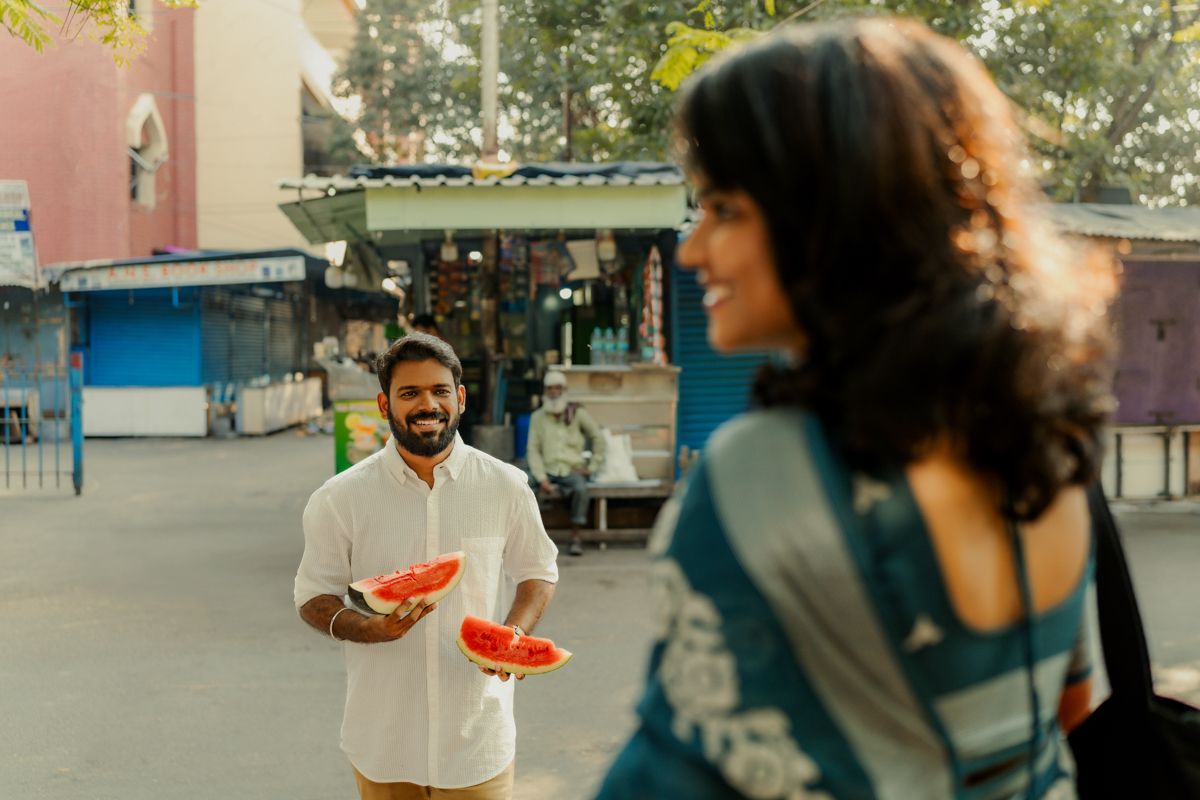 Two people interact on a street with street vendors and storefronts visible in the background.