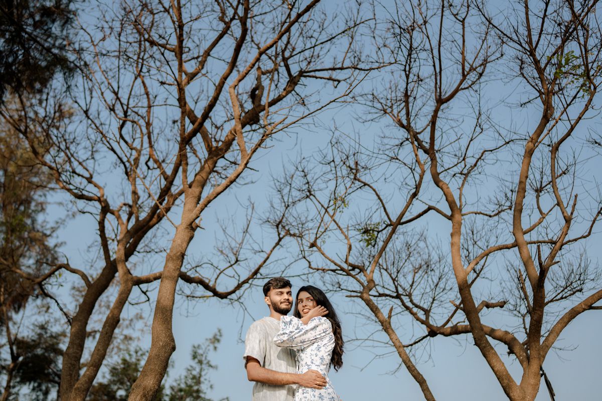 Couple embraces outdoors beneath bare tree branches against bright blue sky on sunny day.