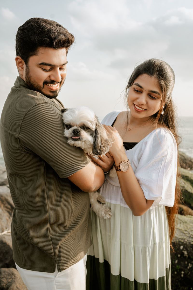 Couple holding small white dog together on rocky coastal setting.