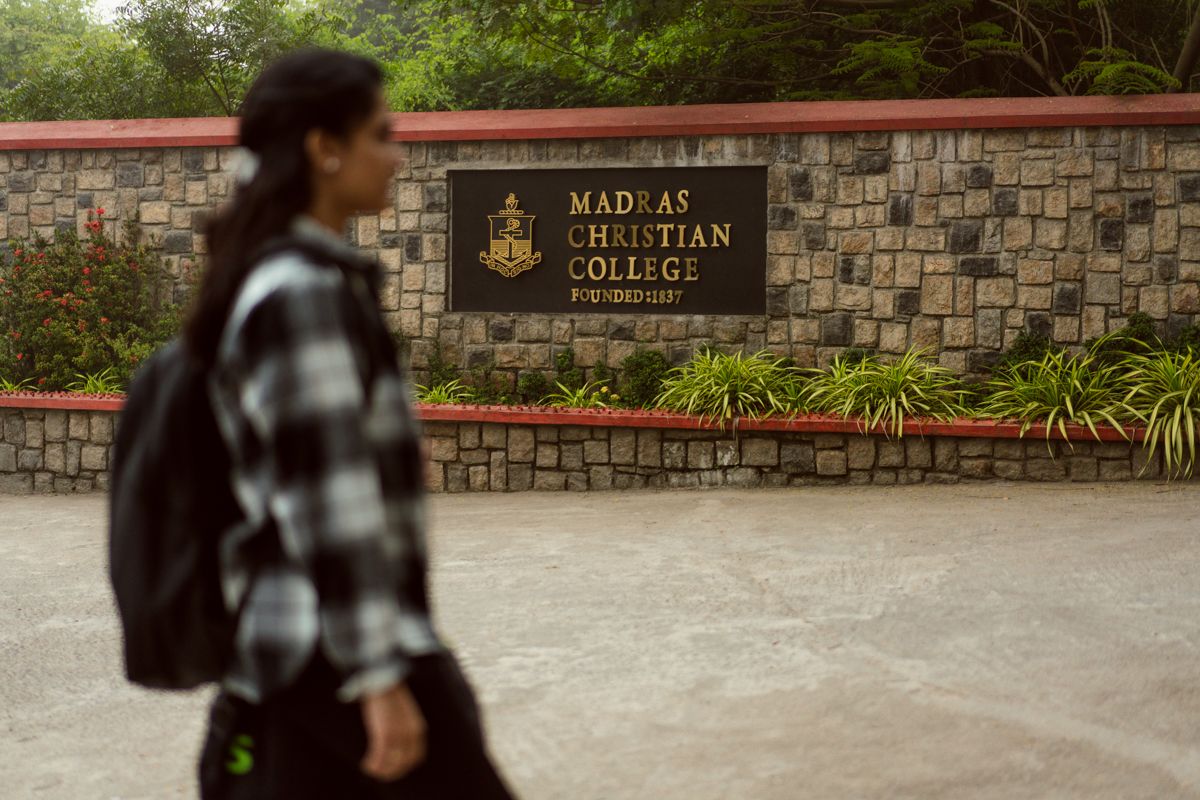 A student walks past the entrance sign of Madras Christian College on a stone wall.