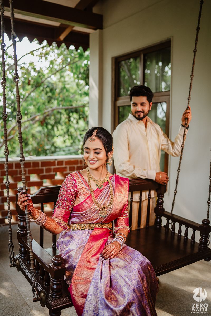 A couple in traditional South Asian attire pose together on a wooden swing bench on a covered porch.